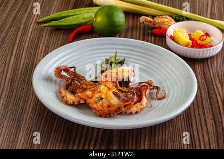 Poulpe grillé servi sur une assiette en céramique texturée avec citron vert, chili rouge, citronnier à feuilles, herbes. Un éclairage chaud met en valeur la dorure grillée et le vert éclatant, re Banque D'Images