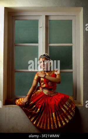 Une danseuse traditionnelle vêtue d'un costume éclatant pose gracieusement près d'une fenêtre. Le cadre met en valeur l'élégance culturelle et l'artisanat, avec des détails complexes Banque D'Images
