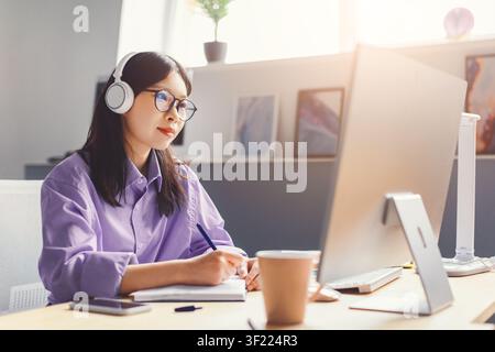 Femme asiatique concentrée portant des écouteurs et travaillant à son bureau à domicile, concept commercial Banque D'Images