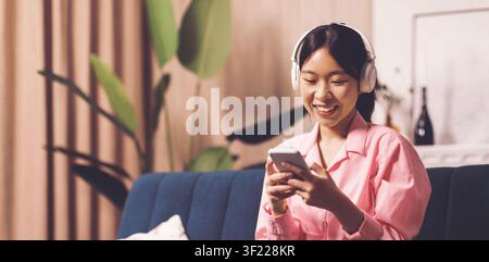 Jeune femme asiatique souriante écoutant de la musique avec des écouteurs tout en utilisant son smartphone à la maison Banque D'Images