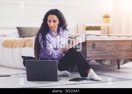 Femme métissée concentrée prenant des notes tout en travaillant depuis chez elle, ordinateur portable devant, concept d'étude Banque D'Images