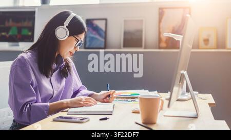 Femme asiatique concentrée portant des écouteurs et travaillant sur un ordinateur portable au bureau à domicile, concept commercial Banque D'Images