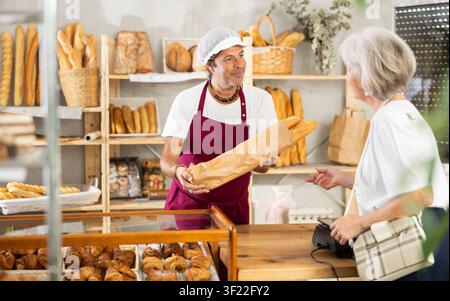 Homme entrepreneur, propriétaire de boulanger met des baguettes dans un sac en papier, sert la cliente senior Banque D'Images