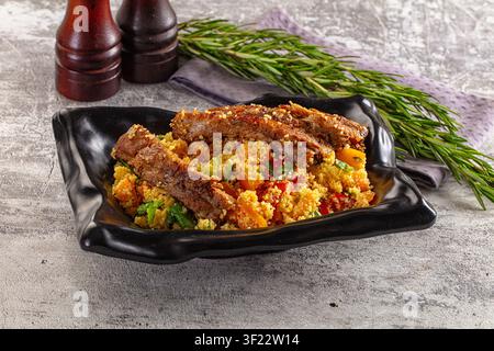 Côtes d'agneau grillées sur un couscous coloré avec des poivrons rouges et jaunes, des herbes fraîches, servies dans un bol en céramique noire. Fond en pierre rustique, brins de romarin Banque D'Images