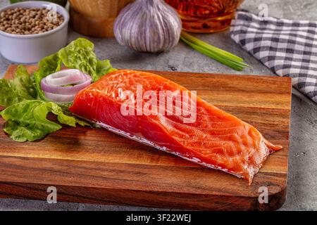 Filet de saumon cru sur planche en bois avec laitue verte et tranches d'oignon rouge. L'arrière-plan comprend de l'ail, des épices, un tissu à carreaux. Un éclairage chaud met en valeur o Banque D'Images