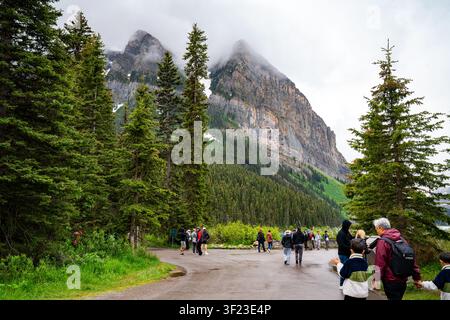 Les visiteurs marchent sous des sommets imposants voilés de nuages près du lac Louise, entourés d'une forêt de feuillus et d'un paysage montagneux spectaculaire dans le parc national de Banff Banque D'Images