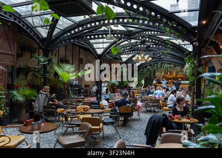Les invités sont assis à des tables dans un café. Certains mangent et boivent tandis que d’autres discutent. Le café est doté de nombreuses plantes et d’un toit vitré. Il est animé et vivant. Banque D'Images
