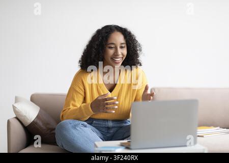 Jeune femme afro-américaine heureuse passant un appel vidéo sur un ordinateur portable assise sur un canapé Banque D'Images
