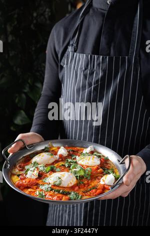 Un chef présente une shakshuka vibrante mettant en vedette des œufs pochés, des herbes fraîches et une sauce tomate riche, parfaite pour un blog culinaire ou un menu de restaurant. Banque D'Images