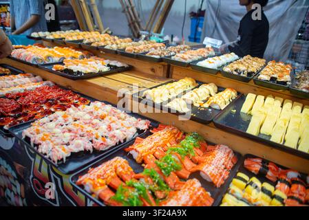 Présentation de plateaux de sushi colorés mettant en vedette une variété de poisson cru, de rouleaux de sushi et de fruits de mer garnis sur un étal de marché vibrant, mettant en valeur des délices japonais frais Banque D'Images