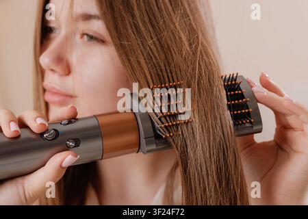 Une jeune femme coiffe ses cheveux avec une brosse-sèche-cheveux, démontrant la facilité d'obtenir des mèches lisses et brillantes à la maison. Banque D'Images