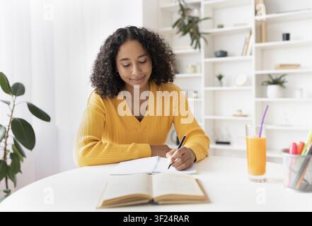 Femme afro-américaine heureuse écrivant dans un cahier à la maison, concept de mode de vie productif Banque D'Images