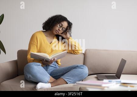 Jeune femme afro-américaine lisant un livre dans un salon confortable, se détendant à la maison Banque D'Images