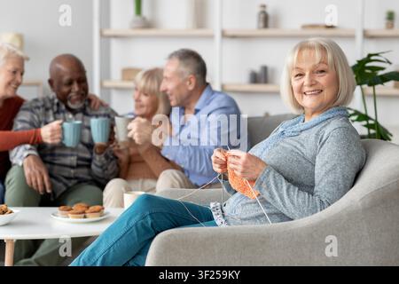 Femme caucasienne âgée souriante tricotant pendant que des amis profitent ensemble à la maison, concept de mode de vie Banque D'Images