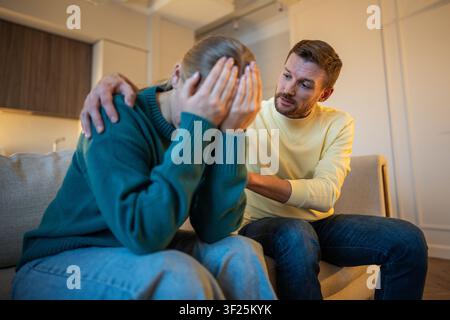 Femme épuisée et frustrée assise sur le canapé, cachant son visage dans ses mains, fatiguée des excuses constantes de son mari Banque D'Images