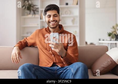 Jeune homme heureux écoutant de la musique avec des écouteurs tout en utilisant son smartphone à la maison Banque D'Images
