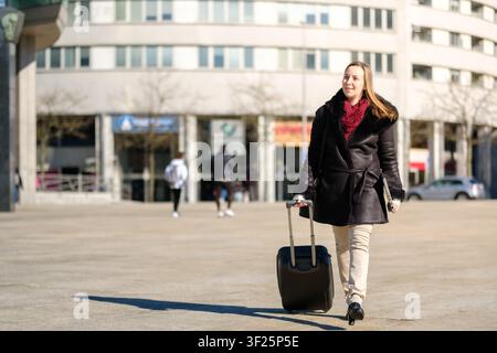 Femme en voyage d'affaires tirant une valise tout en marchant sur une place de ville Banque D'Images