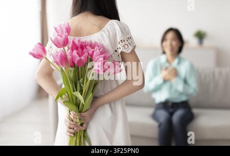 Femme tenant des tulipes roses derrière le dos surprenant une autre femme à la maison, non identifiable Banque D'Images
