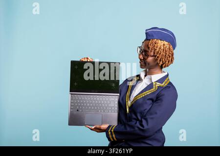 Hôtesse de l'air élégante tenant un ordinateur portable avec un écran vert factice devant un fond bleu. Membre professionnel du personnel aérien présentant un appareil numérique pour des concepts d'affaires, de voyage et de technologie. Banque D'Images