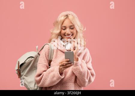 Jeune femme caucasienne souriante utilisant un smartphone dans une tenue confortable, fond rose Banque D'Images
