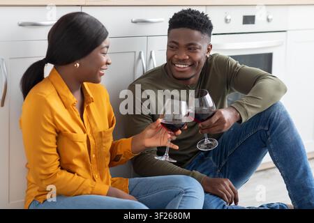 Couple noir heureux portant un toast avec des verres de vin rouge dans une cuisine moderne, concept de convivialité Banque D'Images
