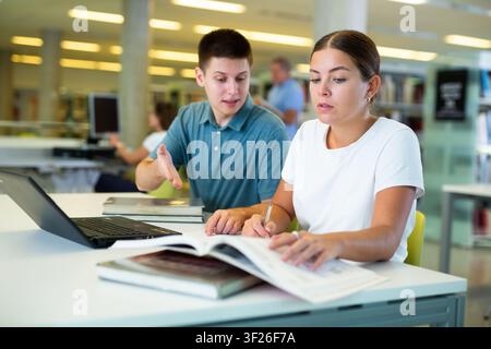 Une jeune femme amicale offre de l'aide à un ami masculin étudiant, trouvant des informations à la bibliothèque Banque D'Images