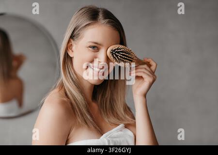 Jeune femme souriante tenant une brosse à cheveux dans la salle de bain, concept bien-être Banque D'Images
