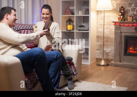 Jeune homme sourit en offrant à sa femme un beau cadeau de Noël sur le canapé, créant un moment d'amour en couple pour les fêtes, avec échange de cadeaux, affection des fêtes et connexion chaleureuse en couple à la maison. Banque D'Images