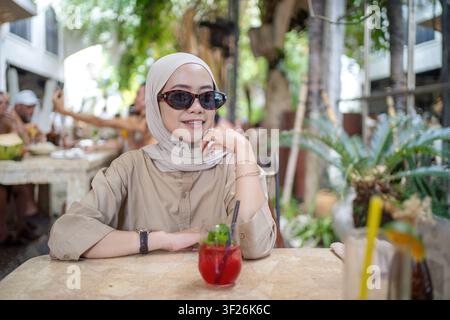 Jeune femme musulmane indonésienne portant des lunettes de soleil et un hijab, assise dans un café en plein air à Bali, Indonésie. Concept de mode, de voyage et de loisirs Banque D'Images