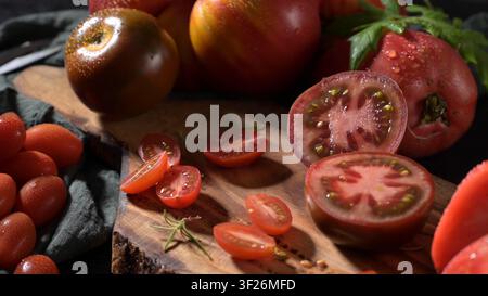 Des tomates anciennes entières et tranchées variées sont disposées sur une planche à découper rustique sombre. Une lumière douce met en valeur la texture de peau rosée et la profondeur vibrante Banque D'Images