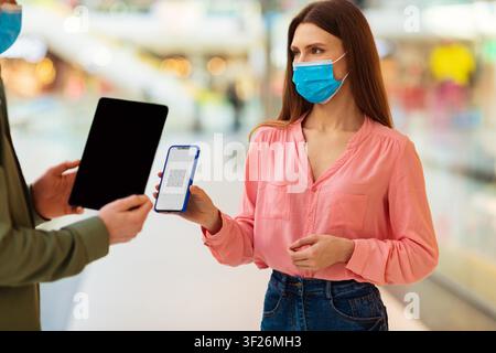 Femme aux cheveux longs tenant un smartphone affichant un code QR dans un centre commercial, portant un masque facial Banque D'Images