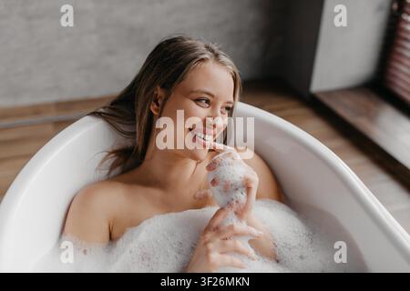 Jeune femme caucasienne souriante profitant d'un bain relaxant avec des bulles dans le spa à domicile Banque D'Images