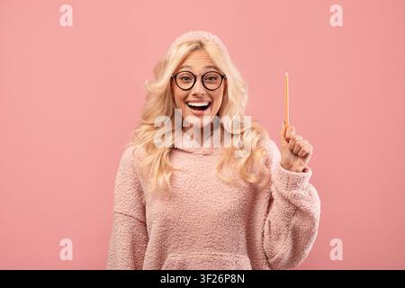 Jeune femme blonde heureuse portant un pull et tenant un crayon, fond rose, concept d'éducation Banque D'Images
