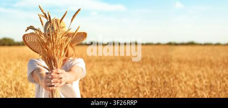 Femme avec chapeau de paille tenant un bouquet d'épis de blé dorés dans un vaste champ agricole sous un ciel bleu, l'arrière-plan présente un vaste paysage baigné de soleil Banque D'Images