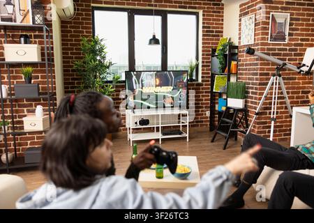Groupe divers jouant ensemble dans le salon alors que des femmes célèbrent leur victoire contre leurs partenaires. L'écran de télévision montre la victoire tandis que des amies femmes taquinent gentiment les hommes pour leur défaite au jeu vidéo. Banque D'Images