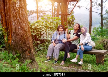 Trois Indonésiennes asiatiques heureuses profitent d'un moment de rire ensemble en étant assises sur un banc en bois dans un parc forestier luxuriant Banque D'Images