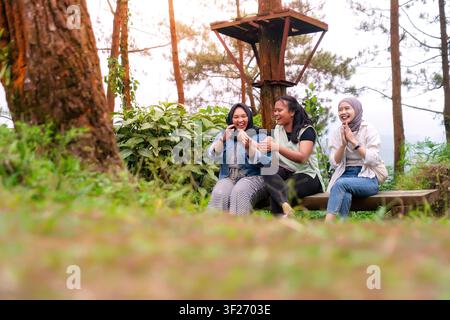 Trois Indonésiennes asiatiques heureuses profitent d'un moment de rire ensemble en étant assises sur un banc en bois dans un parc forestier luxuriant Banque D'Images