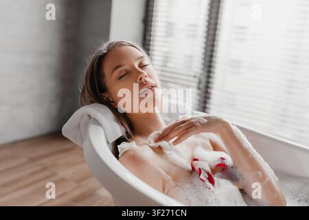 Jeune femme détendue profitant d'un bain moussant avec des pétales de fleurs, concept de spa à domicile Banque D'Images