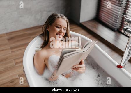 Jeune femme se détendant dans la baignoire avec un livre, profitant d'une expérience spa à la maison Banque D'Images
