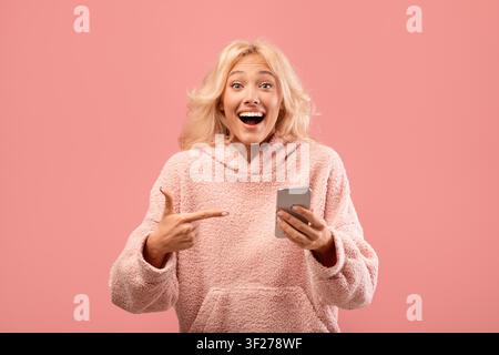 Jeune femme heureuse pointant son smartphone avec excitation, fond rose Banque D'Images