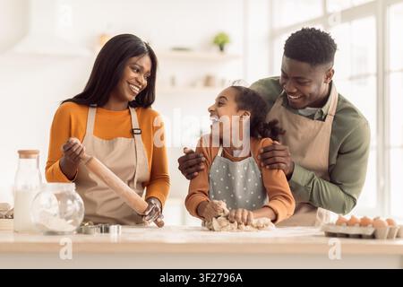 Famille noire heureuse cuisinant ensemble dans une cuisine lumineuse, mère père et fille riant Banque D'Images