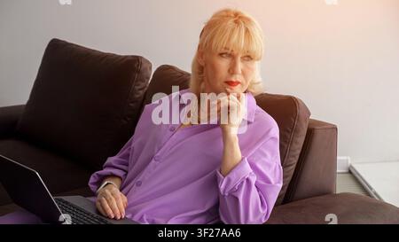 Femme mûre aux cheveux blonds reposant son menton sur sa main, assise sur un canapé avec un ordinateur portable, planifiant pensivement et travaillant à distance depuis un endroit confortable et douillet Banque D'Images