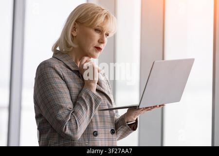 Femme d'affaires âgée debout tenant un ordinateur portable, travaillant et se concentrant sur l'écran, réfléchissant au contenu avec une expression réfléchie dans un moderne Banque D'Images