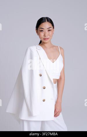 Jeune femme asiatique élégante en costume blanc en portrait vertical avec un regard doux dans un studio lumineux. Convient pour mannequin de mode, beauté et marque personnelle Banque D'Images