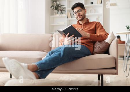 Jeune homme concentré lisant un livre à la maison, se détendant sur un canapé dans un salon moderne Banque D'Images