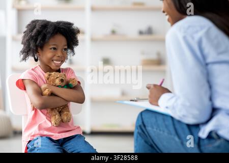 Jeune fille afro-américaine souriante tenant un ours en peluche lors d'une séance de thérapie, concept de psychologie infantile Banque D'Images