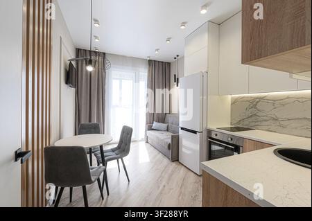 appartement studio moderne et compact avec une salle à manger, une cuisine et un espace de vie avec un canapé près de grandes fenêtres. La palette de couleurs est neutre et c Banque D'Images