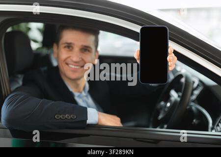 Homme caucasien souriant montrant un smartphone avec écran vide dans une voiture, concept d'affaires Banque D'Images