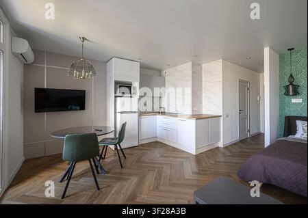 appartement studio moderne comportant une petite cuisine, un coin repas avec des chaises vertes et un lit dans un espace cloisonné. Le design intérieur est épuré et f Banque D'Images