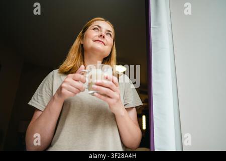 Femme tenant une boisson chaude tout en regardant par la fenêtre Banque D'Images